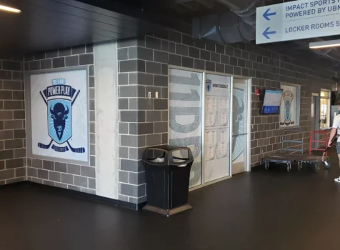 Interior wall and window graphics installed by Ninja Graphics Inc. at an ice hockey facility for the 11 Day Power Play event. The design features a large logo with crossed hockey sticks and a buffalo head on the left wall, along with frosted vinyl treatments on the glass panels displaying event schedules and sponsor information. The space has an industrial-style design with exposed ductwork, brick-patterned walls, and black flooring. Overhead signage directs visitors to locker rooms, while a mounted screen provides additional event details. A few individuals and utility carts are visible in the background.