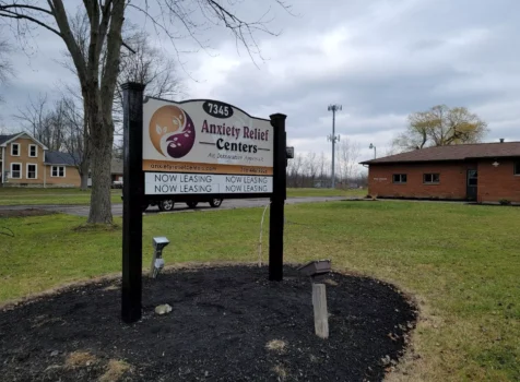 A business sign for Anxiety Relief Centers, featuring a modern, two-tone design with a circular logo that incorporates calming swirls in purple and gold. The sign is mounted on a black wooden frame and displays the business name and the tagline “An Integrative Approach”. Below, there are two lines of text indicating “NOW LEASING” with contact information for the center, including the website (anxietyreliefcenters.com) and phone number (716.446.6226). The sign stands in a grassy area with a suburban backdrop, including a brick building and a cell tower. The sky is cloudy with a hint of overcast.