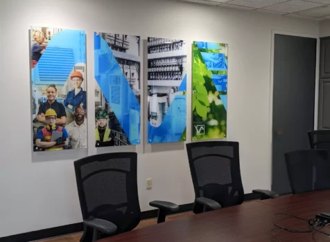 A modern conference room featuring a long wooden table with black mesh office chairs. On the wall, there are four acrylic wall-mounted prints secured with architectural standoffs. The prints display a combination of images, including industrial workers, technical equipment, blue geometric designs, and green leaves, likely representing themes of industry, technology, and sustainability. A door and cabinets are visible in the background.