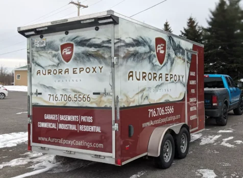 A custom-wrapped trailer for Aurora Epoxy Coatings, featuring a high-end marbled epoxy design in white, gray, and black with burgundy accents. The company’s logo, a stylized “AE” shield, is prominently displayed along with the business name in bold, elegant lettering. The wrap includes contact information (716.706.5566) and a website (www.AuroraEpoxyCoatings.com), as well as a list of services such as garages, basements, patios, commercial, industrial, and residential epoxy flooring solutions. The trailer is parked outdoors on a winter day, with a blue and black pickup truck in the background.