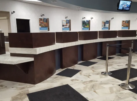 A modern bank interior featuring a curved teller counter with a dark brown base and white marble countertop. The teller stations are evenly spaced, each with privacy dividers. The floor is covered with polished marble tiles, complemented by black anti-fatigue mats in front of each teller station. The walls have a subtle patterned wallpaper, with framed informational posters mounted above the counter. Small security cameras are installed along the wall for surveillance. A digital screen is mounted near the ceiling, displaying content for customers. A retractable queue barrier with stainless steel posts guides customers in line. The overall design is clean, professional, and inviting.