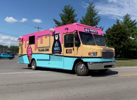 A vibrant and colorful ice cream truck for Big Papa’s Creamery featuring a custom vinyl wrap. The design resembles an ice cream cone, with a waffle-textured beige lower half, a bright blue bottom, and a dripping pink ice cream effect on the roof. The wrap includes playful illustrations of ice cream cones, the Big Papa’s Creamery logo with a bear mascot, and the business’s contact information (716.860.2185) along with social media icons. The truck is a large Freightliner step van, parked on a sunny day with a bright blue sky, scattered clouds, and green trees in the background.