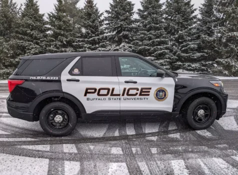 A Buffalo State University Police vehicle featuring a custom black and white vinyl wrap parked on a snowy day. The vehicle is a black Ford Police Interceptor SUV with white doors displaying bold “POLICE” lettering in bronze and black, along with the Buffalo State University name and the official New York State University Police emblem. The department’s contact number (716.878.6333) is printed on the rear side panel. Snow is falling, and the ground is covered with light snowfall, with tire tracks visible. In the background, a row of evergreen trees dusted with snow adds to the winter setting.