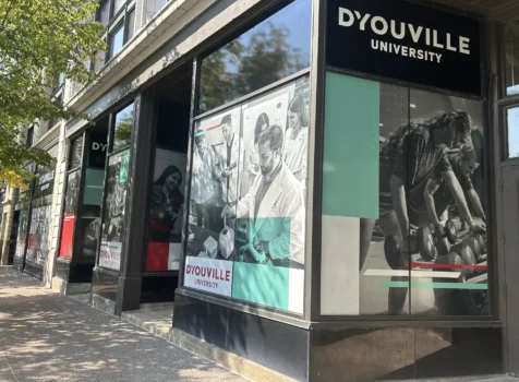 A storefront-style exterior of D’Youville University, featuring large promotional window graphics displaying students engaged in various academic and athletic activities. The black, white, and teal-colored visuals include images of students in lab coats conducting experiments and individuals working out with dumbbells.