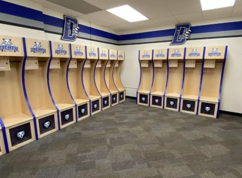 A clean and organized locker room featuring custom wooden lockers for Daemen Athletics. Each locker has a curved design with blue trim, a Daemen Athletics logo at the top, and a Wildcats mascot emblem on the lower storage compartment.