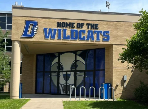 Exterior signage for Daemen University featuring bold, dimensional lettering reading ‘Home of the Wildcats’ above the entrance. The large ‘D’ logo with claw marks is prominently displayed. The glass entrance doors and windows are wrapped with a striking graphic of a wildcat mascot. This custom signage and window graphics were designed and installed by Ninja Graphics Inc.