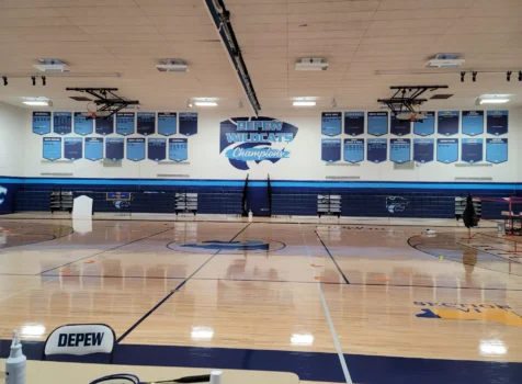 A gymnasium for the Depew Wildcats, featuring a polished wooden basketball court with blue and white markings. The back wall displays “DEPEW WILDCATS Champions” with multiple blue championship banners recognizing the school’s athletic achievements in various sports.