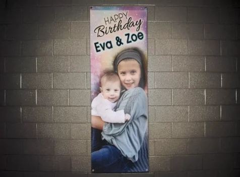 A vertical birthday banner hangs on a dark brick wall. The banner features a colorful, starry background with the words “Happy Birthday Eva & Zoe” in a playful, decorative font at the top. Below the text, there is a photograph of two young children—an older girl with a headband and a younger baby—both smiling while the older girl embraces the baby. The banner is secured with metal grommets at the corners.