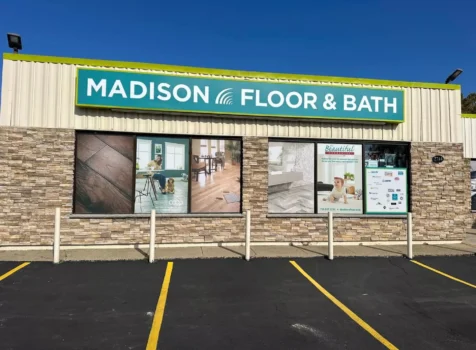 A storefront of “Madison Floor & Bath” is shown on a sunny day. The building has a combination of beige metal siding and a stone facade. A large green and white sign with the store’s name is mounted above the windows. The windows feature promotional images of flooring, a modern bathroom, and a baby, along with a list of brands and services. The parking lot in front has yellow-lined spaces, and white bollards are positioned near the entrance. The sky is clear and blue.