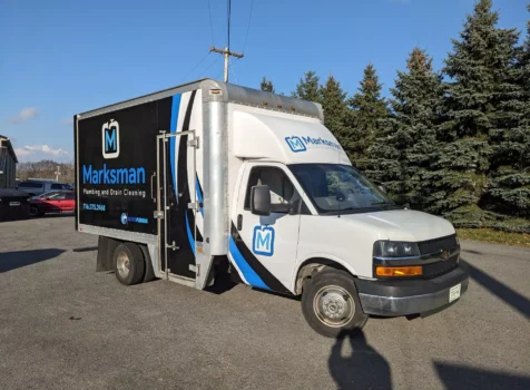 A Marksman Plumbing and Drain Cleaning service truck with a custom vehicle wrap parked outdoors on a sunny day. The truck is a white Chevrolet box truck with a sleek black, blue, and white vinyl wrap featuring the Marksman Plumbing logo, contact information, and branding. The wrap design includes dynamic blue and white swooping lines that complement the company’s logo. The truck is parked on a paved lot with evergreen trees and a clear blue sky in the background. The reflection of the photographer is visible in the lower right corner of the image.