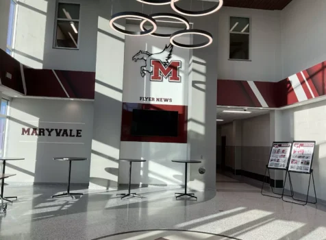 A modern and well-lit entryway to the Maryvale Flyers gymnasium. The space features high ceilings, large windows allowing natural light to stream in, and a clean, polished floor. A large “M” logo with a flying horse is displayed prominently above a digital screen labeled “FLYER NEWS.” The walls incorporate Maryvale’s maroon and white color scheme with dynamic diagonal stripe accents.