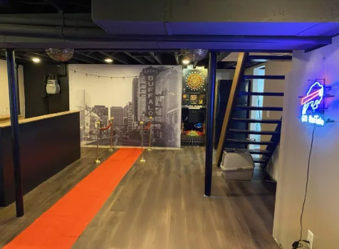 A stylish basement entertainment area featuring a custom vinyl wall graphic of downtown Buffalo, installed by Ninja Graphics Inc. The space includes a red carpet with gold stanchions leading to the wall mural, a wooden bar with a black base, a dartboard game, and a staircase with modern open steps. A neon Buffalo Bills sign illuminates the right wall, adding a sports-themed accent. The ceiling is painted black with exposed beams and disco balls, creating a sleek and modern ambiance.