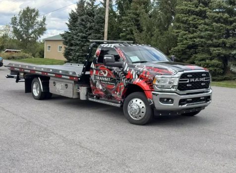 A Transit Towing & Recovery flatbed tow truck with a bold custom wrap is parked on a paved road with trees and a small building in the background. The truck is a heavy-duty RAM model with chrome accents and a rugged design. The wrap features a red, black, and gray graphic pattern with the company’s logo, which includes a stylized eye and the words “Transit Towing & Recovery.”