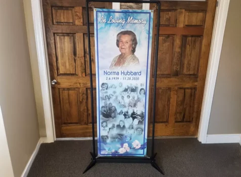 A memorial banner honoring Norma Hubbard (2.6.1939 - 11.28.2020), displayed on a black metal stand in front of wooden double doors. The banner has a blue and white color scheme with the words “In Loving Memory” at the top. It features a large portrait of Norma Hubbard in the upper section, while the lower section contains a collage of black-and-white family photos. The bottom of the banner is adorned with floral and butterfly graphics, creating a soft and elegant tribute. The display is set in a well-lit indoor space with beige walls and a carpeted floor.