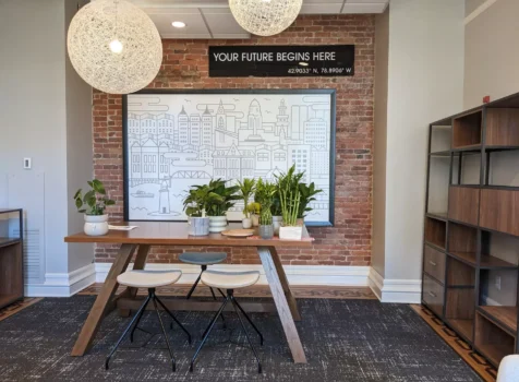 A modern office space with an inviting and creative atmosphere. The focal point is a wooden table with black metal stools, surrounded by potted green plants. Above the table, two spherical pendant lights made of woven material hang, casting soft illumination. The background features an exposed brick wall with a large framed line-art illustration of a cityscape. Above the artwork, a black sign reads “YOUR FUTURE BEGINS HERE” with coordinates displayed below. To the right, a tall open shelving unit with wood and metal elements provides storage. The space combines industrial, contemporary, and natural design elements for a warm and inspiring workspace.