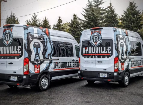 Two D’Youville Saints team vans with custom wraps are parked side by side in an outdoor setting with trees in the background. The wraps feature a bold design with a large, stylized Saint Bernard dog mascot in blue, white, and black, along with the “D’Youville Saints” logo in red, white, and black.