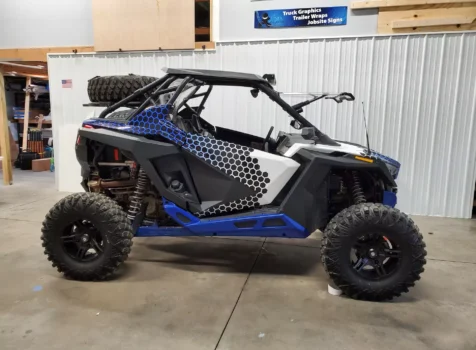 A customized off-road side-by-side vehicle with a blue, black, and white wrap featuring a hexagonal pattern is parked inside a workshop.