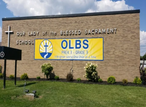 The exterior of Our Lady of the Blessed Sacrament School, a brick building with large silver lettering displaying the school’s name and a white cross mounted on the left side. A bright yellow and blue banner is mounted on the wall, promoting OLBS for Pre-K 3 to Grade 3 with the tagline “A Brighter World One Child at a Time.” The banner features a stylized logo of a cross with sun rays. The school grounds have neatly trimmed grass, small bushes, and flowering plants. A black marquee sign stands in the foreground, and the sky is partly cloudy with blue skies visible.