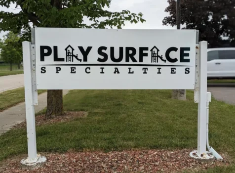 An outdoor business sign for Play Surface Specialties, mounted on a white metal frame with a gravel base. The sign features bold black lettering, with the words “PLAY SURFACE” creatively incorporating playground slide icons into the letters “A” to represent children playing. Below, the word “SPECIALTIES” is displayed in a smaller, evenly spaced font. The sign is set against a backdrop of green grass, a tree, and a parked white van in a suburban area.