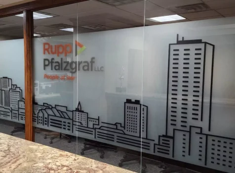 Frosted glass office partition featuring a custom vinyl design by Ninja Graphics Inc. for Rupp Pfalzgraf LLC. The glass showcases a modern black city skyline graphic along the lower half, while the firm’s logo and tagline, “People at law,” are displayed in the upper section. The sleek, professional design enhances privacy while maintaining an open office feel. A granite countertop is visible in the foreground, with office chairs and a workspace behind the glass partition.