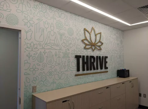 An interior wellness space featuring a custom-designed wall with a light green pattern of yoga, meditation, and nature-inspired line art. The focal point is a three-dimensional gold and black wall sign reading “THRIVE,” accompanied by a stylized lotus flower emblem above it. Below the sign, there is a light wood cabinet with multiple drawers, and a small black electronic device sits on top. The space has a modern, minimalist aesthetic with recessed lighting and a clean, calming atmosphere.