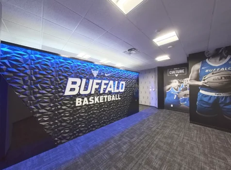 A modern college basketball locker room features a bold “BUFFALO BASKETBALL” sign on a textured black wall illuminated by blue lighting.