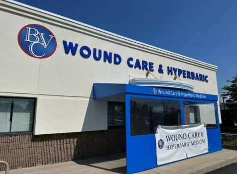 A medical facility with a sign reading “Wound Care & Hyperbaric” in bold blue letters is displayed on the exterior of a white and beige building. A circular logo with the letters “BVC” is positioned to the left of the text. The entrance features a blue awning with “Wound Care & Hyperbaric Medicine” printed on it. Below the awning, a temporary white banner with the same text and logo is attached. The building has a combination of smooth white upper walls and textured brown lower walls. The sky is clear and blue.